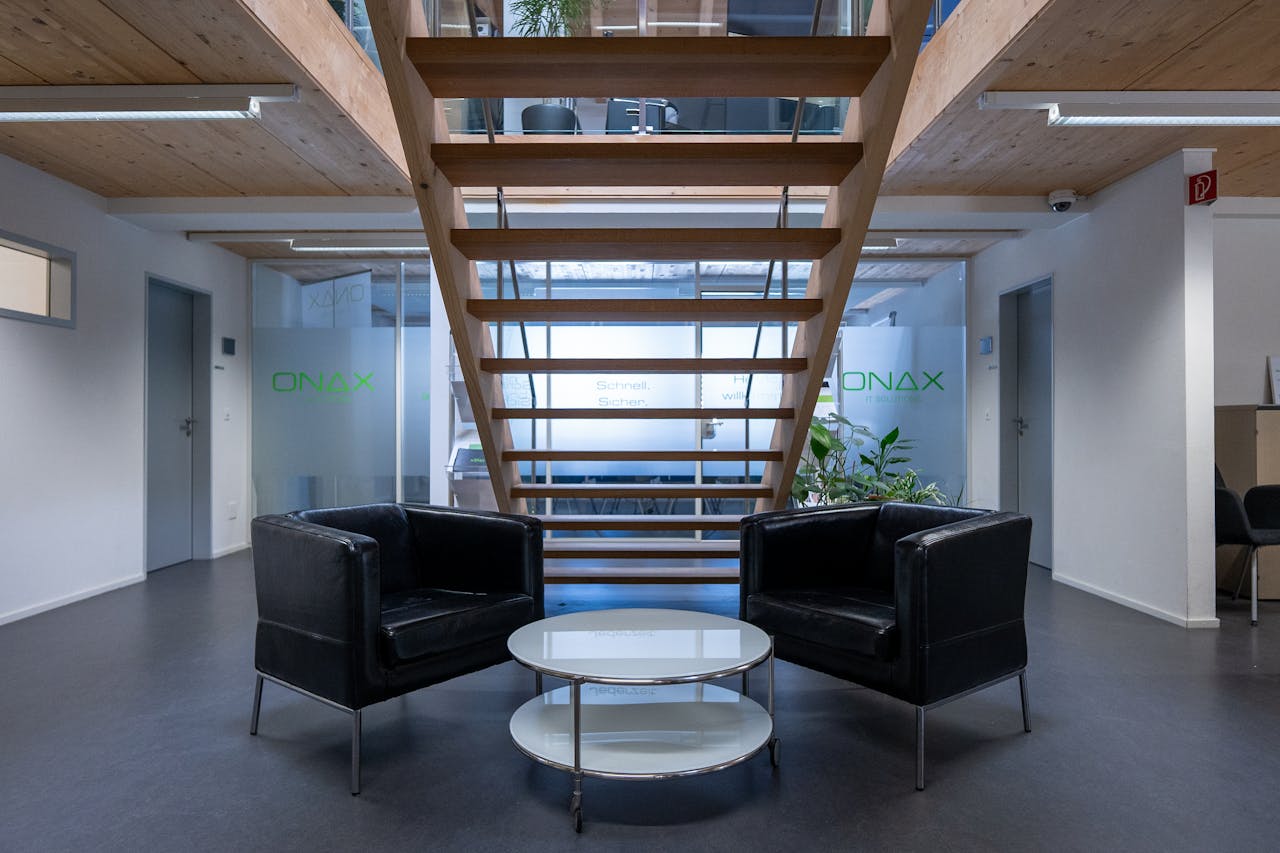 Contemporary office lounge in Cazis with sleek black chairs and a glass table under a wooden staircase.